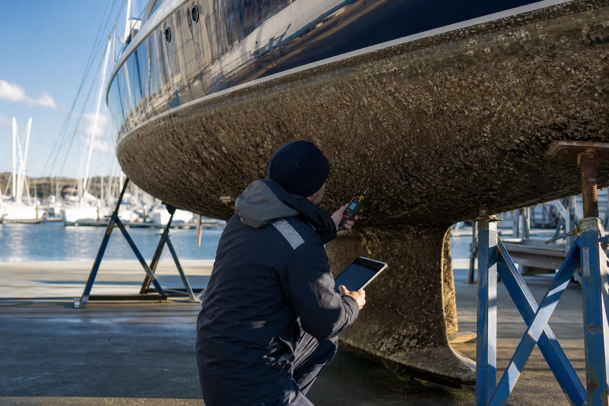 Un expert maritime relevant l&rsquo;état des œuvres vives d&rsquo;un voilier au sec, tablette et humidimètre en main